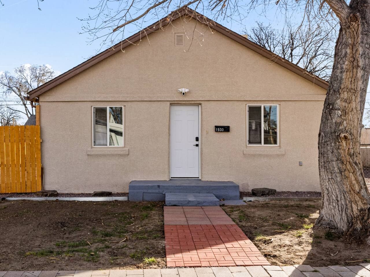 A single-story house with a front door, two windows, a small porch, and a tree in the yard.