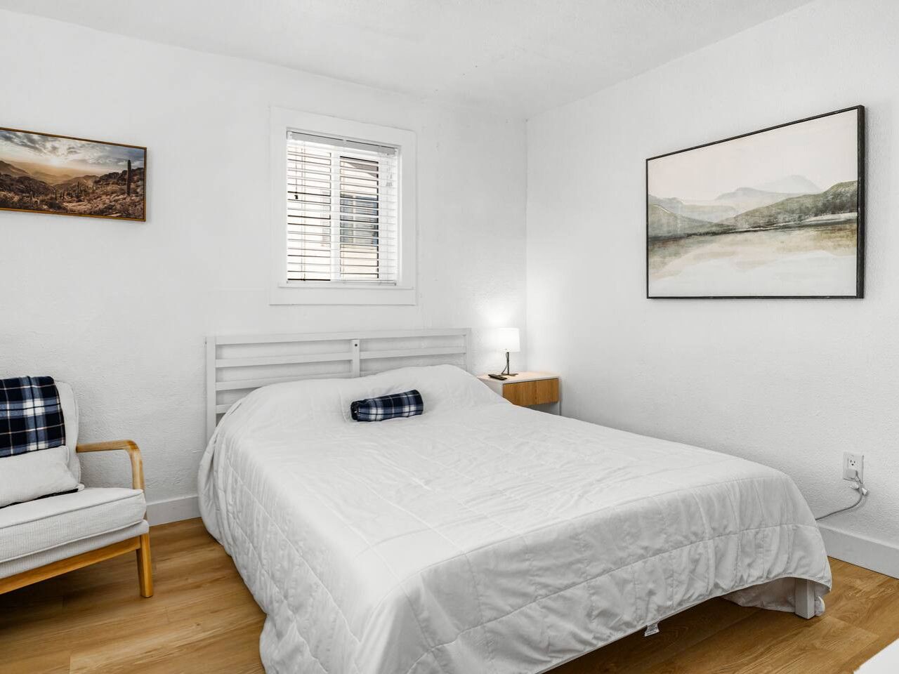 Een slaapkamer met een bed, een nachtkastje, een stoel, een raam met jaloezieën, en twee wanddecoraties.