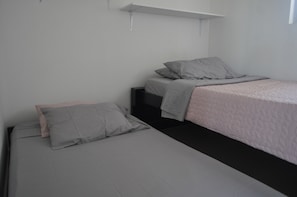 A single bed with a grey mattress and pillows, a bedside table, and a white wall with a shelf. - Aqua blue (Santa Marta)