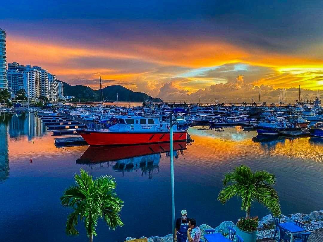 Un puerto deportivo con barcos amarrados, un barco rojo al frente y edificios al fondo.