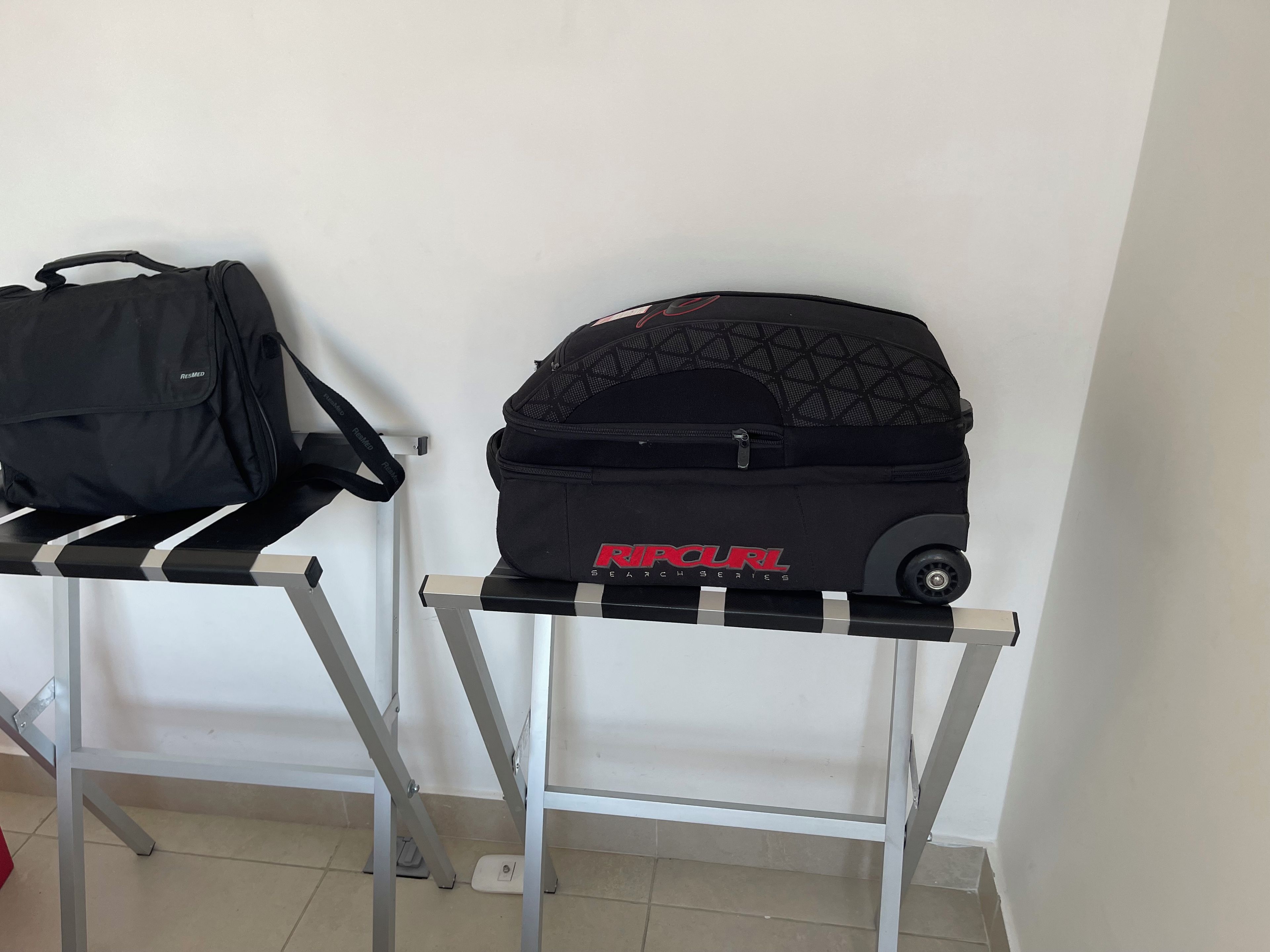 A black bag and a black wheeled bag on a metal table against a white wall.