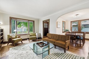 A spacious living room with a glass coffee table, a sofa, a dining table with chairs, and large windows offering a view of the outdoors. - Urban Oasis w/ 10 beds•Sauna•Solar•Wellness Studio•Fenced Yard•Garage (Falcon Heights)