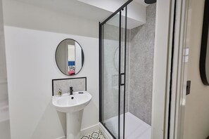 Un baño moderno con un lavamanos blanco, un espejo redondo y una zona de ducha con mampara de vidrio.