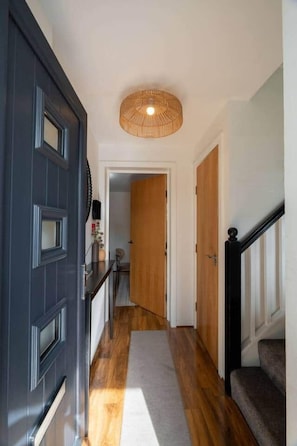 A hallway with a dark door, a light-colored wall, a wooden floor, and a ceiling light fixture. - Lochburn House | Garden, Parking & Stylish 3BR (Glasgow)