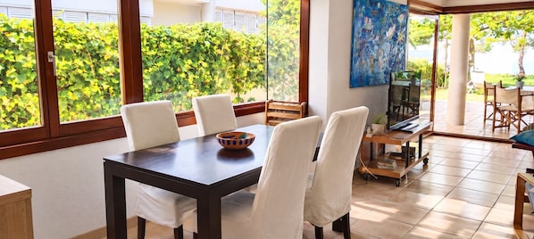 A dining area with a table and chairs, a television, and a view of greenery outside.