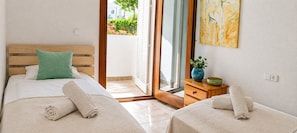 Habitación de hotel con dos camas, cabecero de madera, almohada verde, un cuadro, una planta y una mesita de noche de madera con un cuenco.