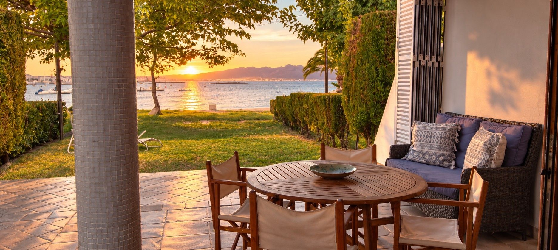 A patio with a round table and chairs, overlooking a body of water with boats and a sunset.