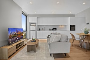 A modern living room with a flat-screen TV, a grey sofa, and a dining area with a round table and chairs. - Balcony 2-bed Apartment Near Albert Park (Melbourne)