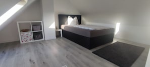 A bedroom with a sloped ceiling, a bed with a dark headboard, a nightstand, a storage unit with floral patterns, and a grey rug. - Vrbo Property (Euskirchen)