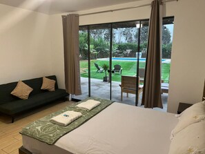 Een hotelkamer met een bed, een slaapbank, een klein tafeltje en uitzicht op een tuin en zwembad.
