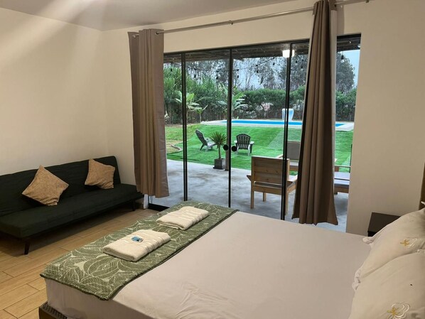 A hotel room with a bed, a sofa, a small table, and a view of a garden and pool.