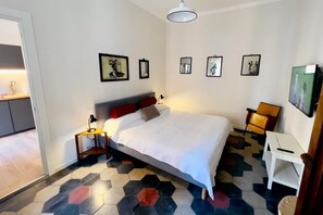 Een slaapkamer met een bed, een bureau en een stoel. Er hangen foto's aan de muur en een deur naar een andere kamer.