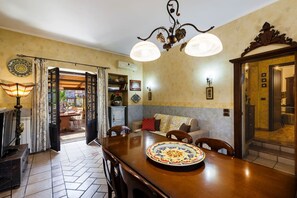 A dining area with a wooden table, chairs, and a decorative plate. There is a sofa with cushions, a chandelier, and a wall-mounted clock. - Villa Rubino - Confortevole Casa con Ampio Spazio (Catania)