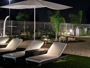A nighttime outdoor seating area with lounge chairs and a large umbrella, surrounded by a white fence and landscaped garden. - Villa Elisia - Villetta con Piscina Privata (Augusta)