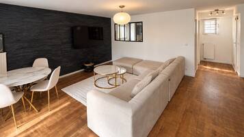 Un salon moderne comprenant un canapé d’angle, une table à manger avec des chaises, une table basse ronde et un téléviseur à écran plat fixé sur un mur foncé.