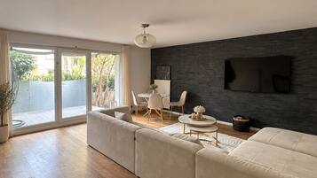 Un salon moderne doté d’un grand canapé d’angle, d’un coin repas avec des chaises, d’un téléviseur à écran plat fixé sur un mur sombre et d’une porte-fenêtre coulissante donnant sur un espace extérieur.