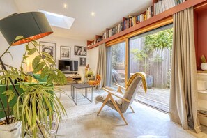 A modern living room with a sofa, a coffee table, a TV, and a bookshelf. There is a sliding glass door leading to a patio with a wooden deck. - Hostcare - Maison unique dans ancien bar rénové (Nantes)