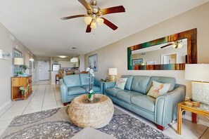 A spacious living room with a blue sofa, a round woven coffee table, and a ceiling fan. - 🌅Walk to Beach | Gulf & Lake Views | Pelican Terrace 905🌴 (Destin)