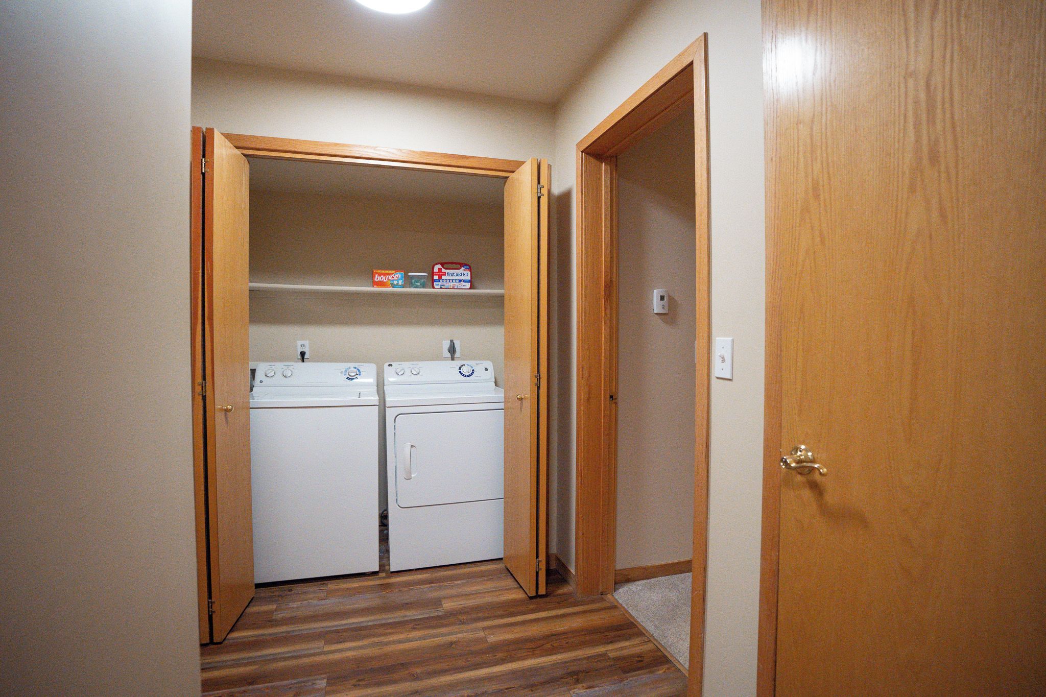A laundry room with two white front-loading washing machines and a shelf with a first aid kit and a box of Bounce detergent.