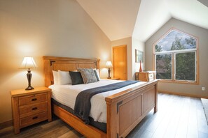 Un dormitorio con una cama de madera, dos mesitas de noche, una mesita auxiliar, un ventanal con vistas y una puerta.