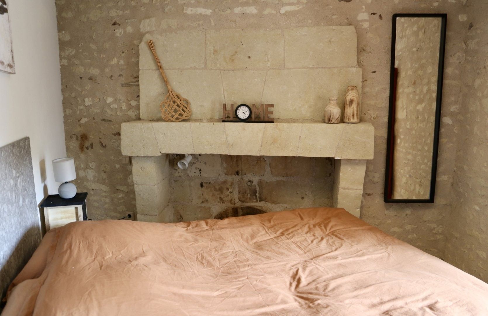 A bedroom with a bed, a nightstand, a wall-mounted shelf with a clock, a woven bag, and a framed mirror.