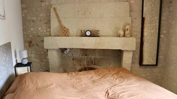A bedroom with a bed, a nightstand, a wall-mounted shelf with a clock, a woven bag, and a framed mirror.