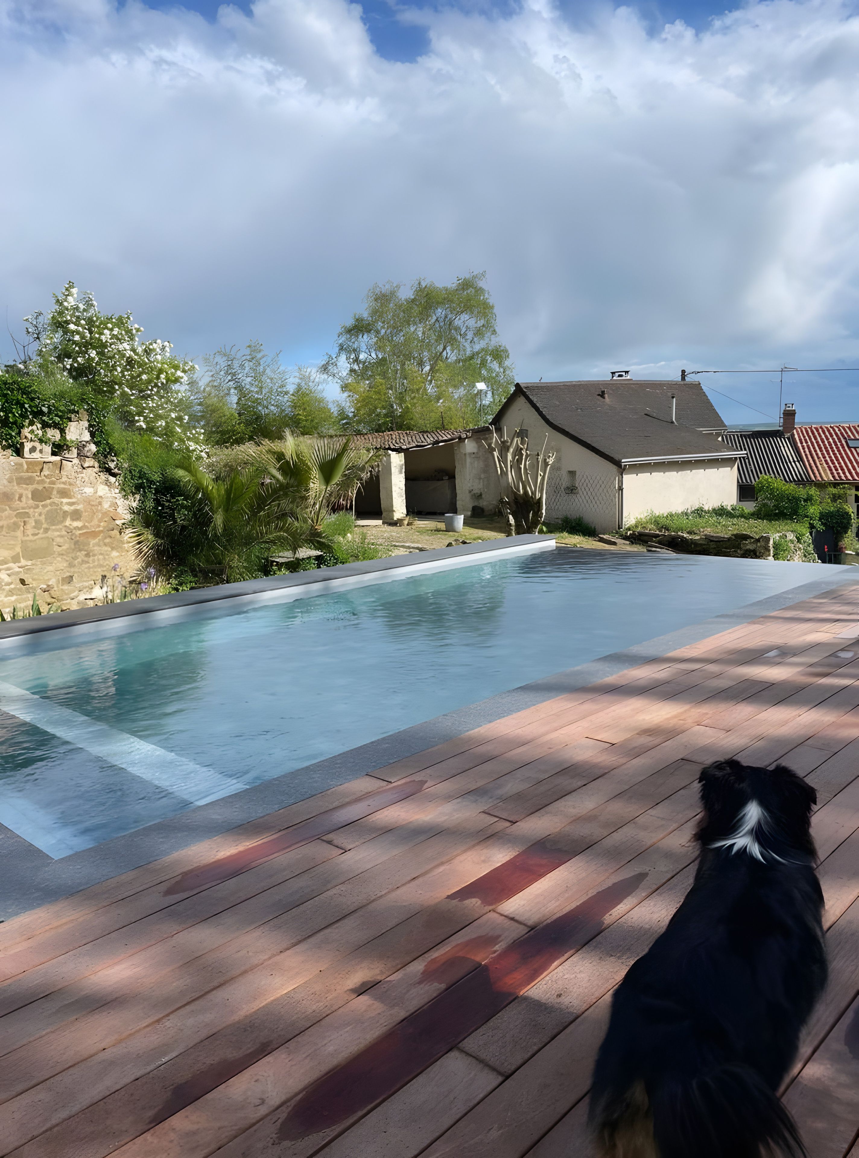 Une piscine dotée d’une terrasse en bois, entourée de verdure et bordée d’un mur de pierre.