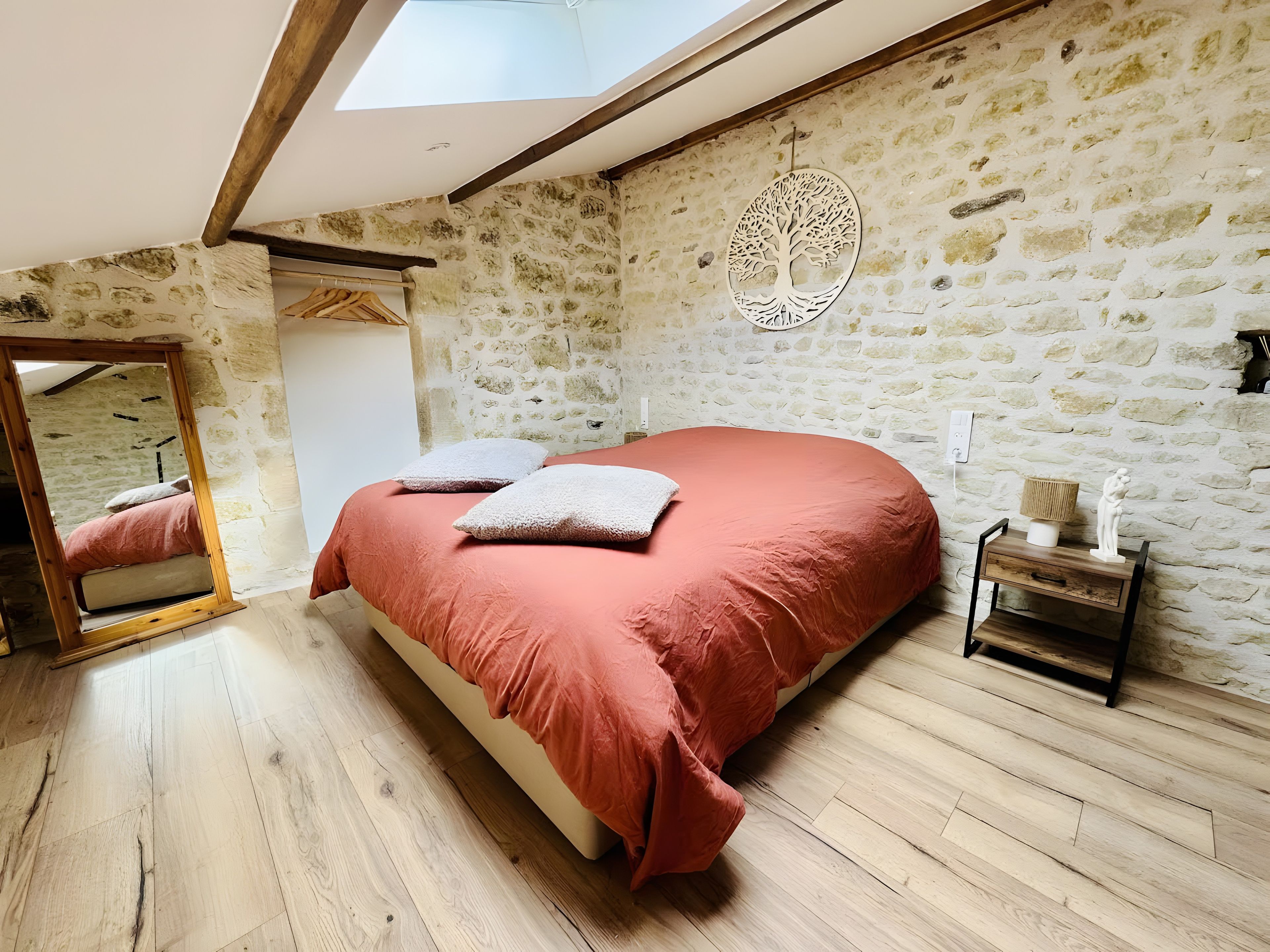 Une chambre avec un mur en pierre, un lit recouvert d’une courtepointe rouge, un plancher en bois et une lucarne.