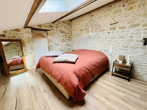 Une chambre avec un mur en pierre, un lit recouvert d’une courtepointe rouge, un plancher en bois et une lucarne.