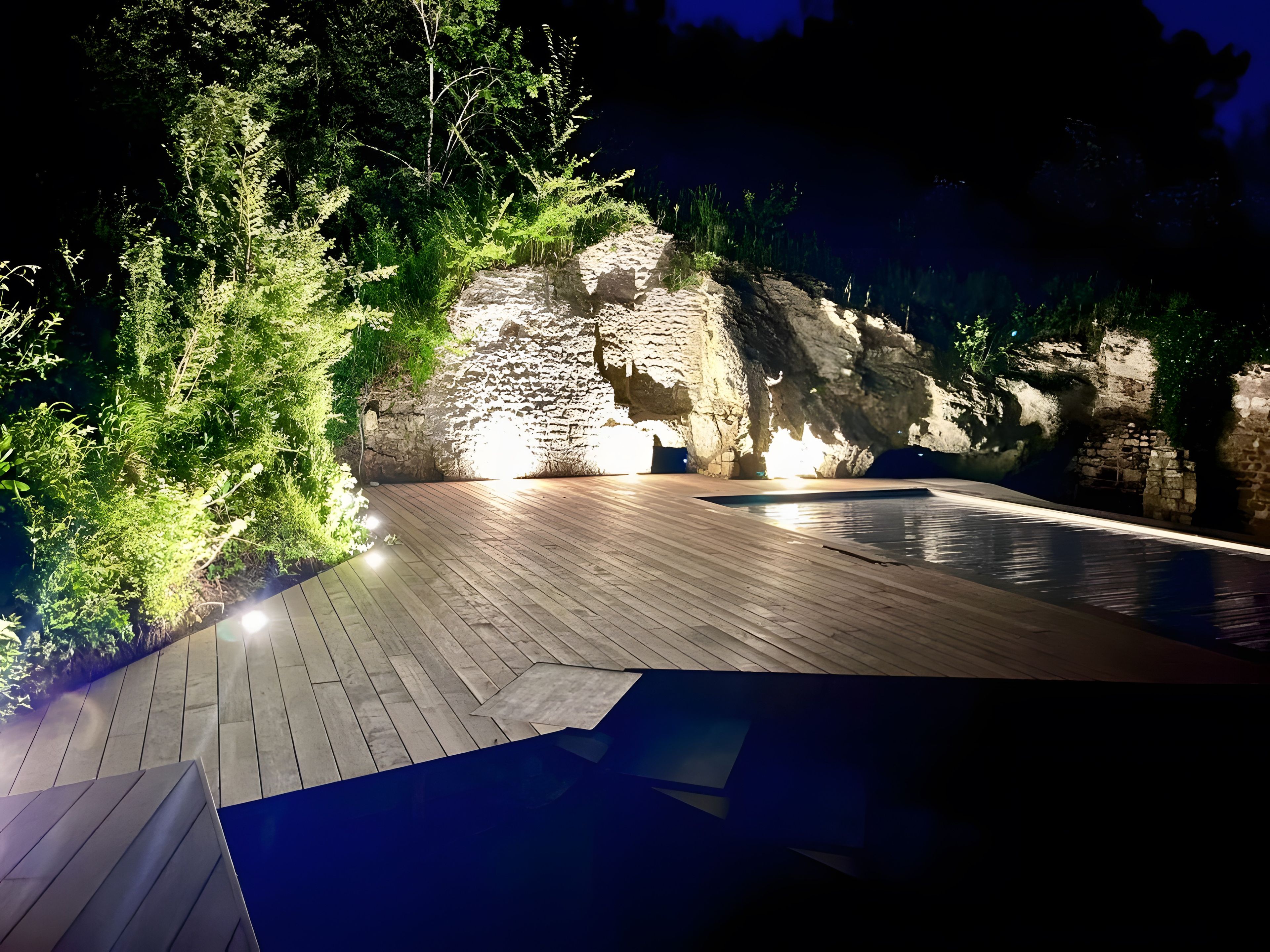 Eine Holzterrasse im Freien mit einem Pool, umgeben von Grünpflanzen und beleuchtet durch künstliche Beleuchtung.
