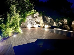 Una terrassa de fusta a l'aire lliure amb piscina, envoltada de vegetació i il·luminada amb llums artificials.