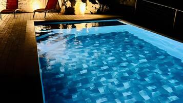 Una piscina con suelo de baldosas, rodeada por una terraza de madera y sillas rojas.