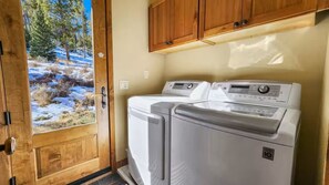 A laundry room with two front-loading washing machines, wooden cabinets above, and a door leading to a snowy outdoor scene. - Serene home on 2 Acres, Minutes to Slopes, Private Hot Tub (Breckenridge)