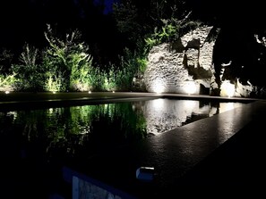Un área de piscina nocturna con muro de piedra y vegetación.