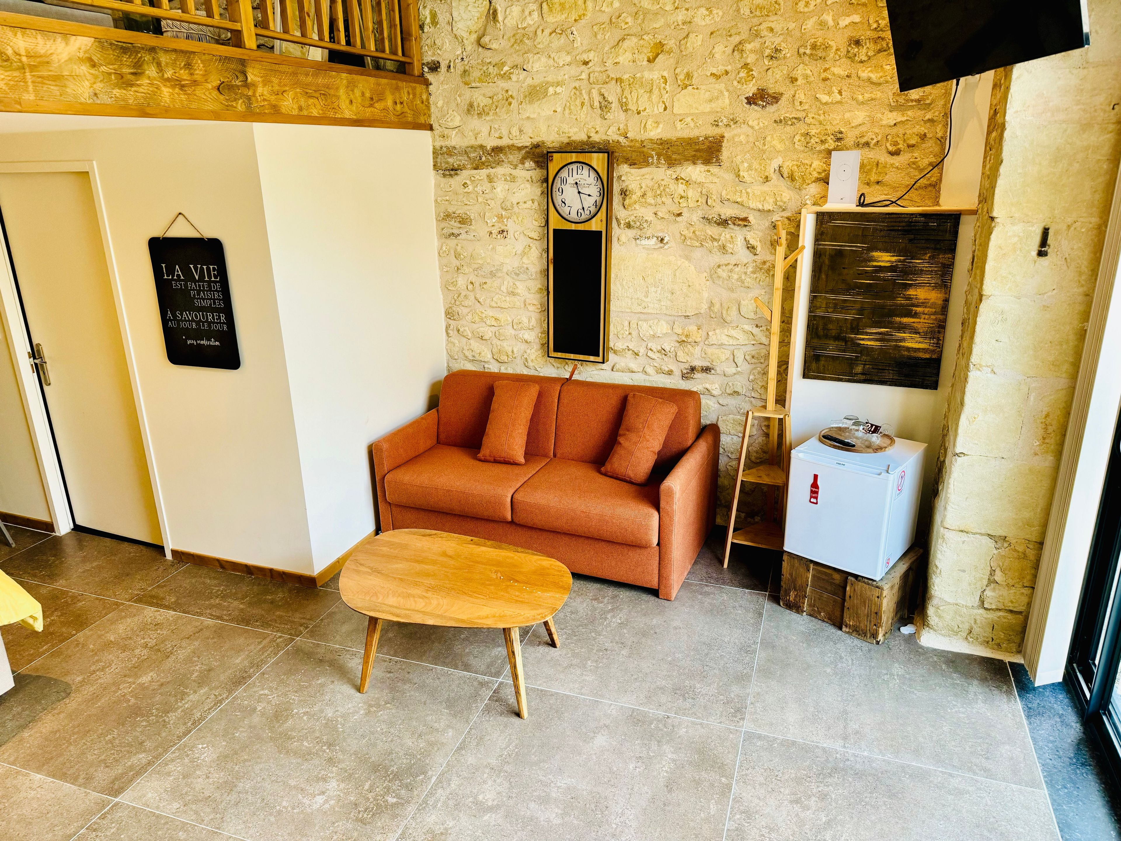 Une pièce chaleureuse avec un mur en pierre, un banc en bois, un canapé, une horloge et un petit réfrigérateur.