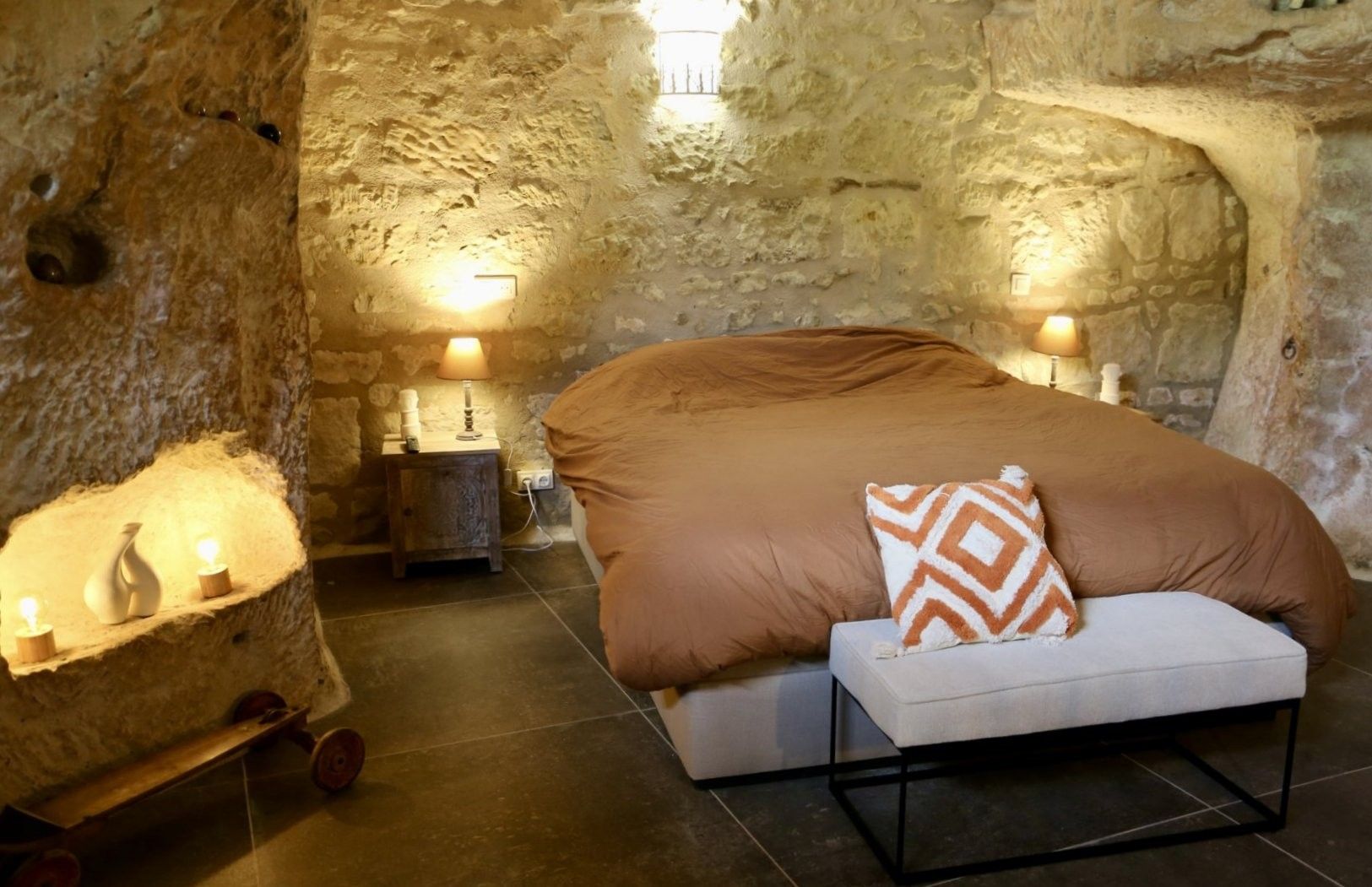 A bedroom with a stone wall, a bed with a patterned pillow, bedside lamps, and a small wooden nightstand.