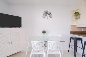 A modern dining area with a glass table and white chairs, a flat-screen TV mounted above, and a decorative wall piece. - ☀️Summer Breeze 302 | Gulf Views~Pool+Hot Tub~Balcony~Walk to Beach⛱️ (Miramar Beach)