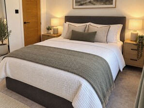 A neatly made bed with a grey blanket, two bedside tables with lamps, a wooden door, and a framed picture on the wall. - Meadow View Cottage (Gloucester)