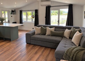 A modern living room with a grey sectional sofa, a small kitchen island, and a view of the outdoors through large windows. - Meadow View Cottage (Gloucester)