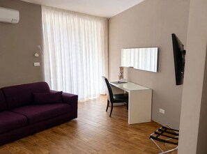 Een kamer met een paarse bank, een wit bureau, een stoel, een aan de muur bevestigde televisie en een raam met witte gordijnen.