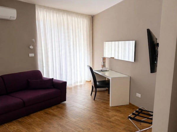 A room with a purple sofa, a white desk, a chair, a TV mounted on the wall, and a window with curtains. - Vrbo Property (Napoli)