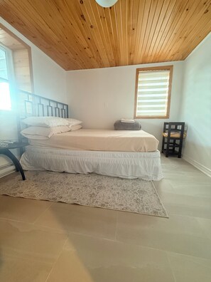 A bedroom with a wooden ceiling, a bed with white bedding, a small table with a chair, and a window with blinds. - Cozy Current Chalet with the backyard facing  Ottawa River (Montebello)
