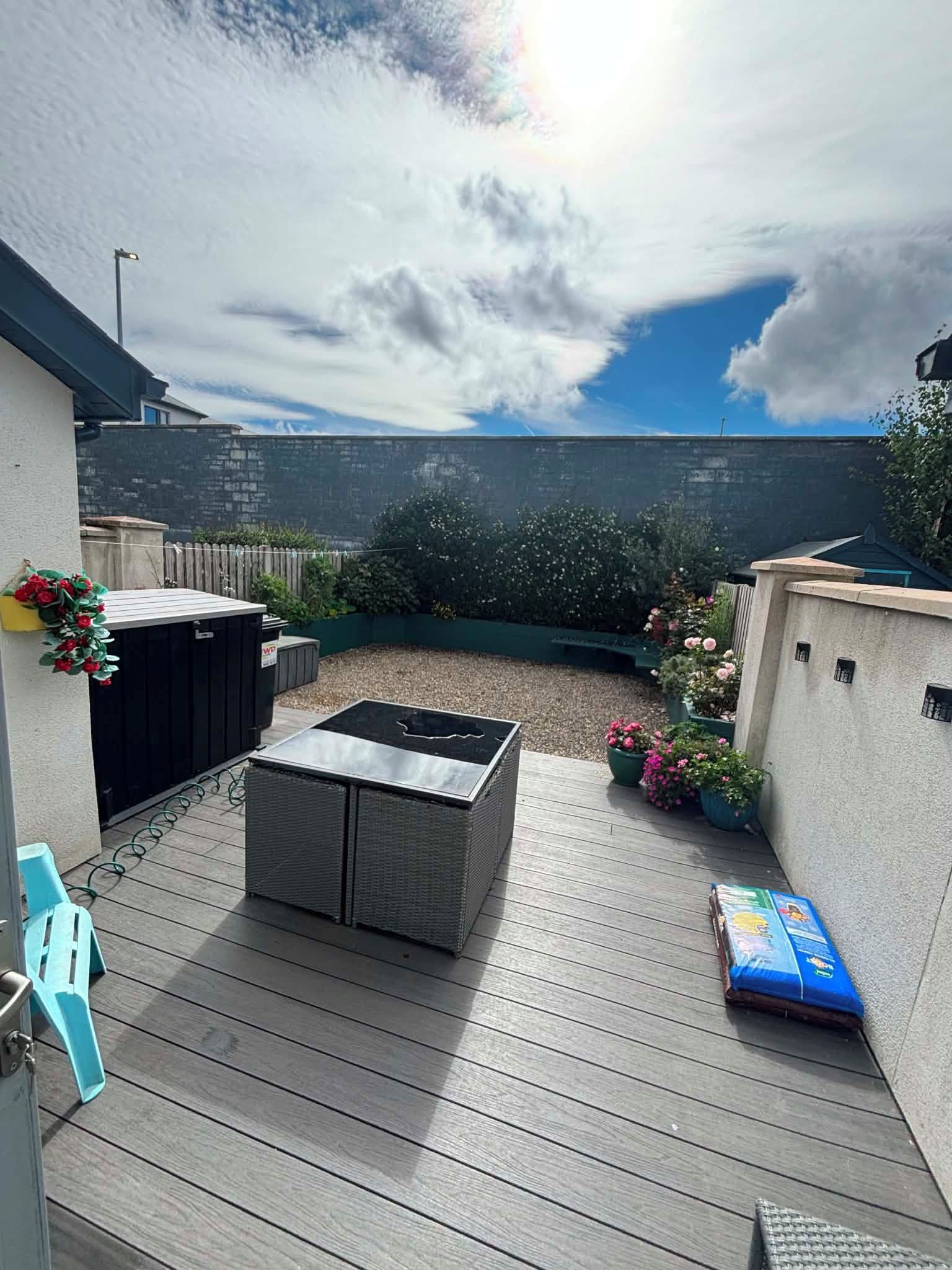 Um deck de madeira com área de estar integrada, um pequeno jardim com flores e um céu limpo.