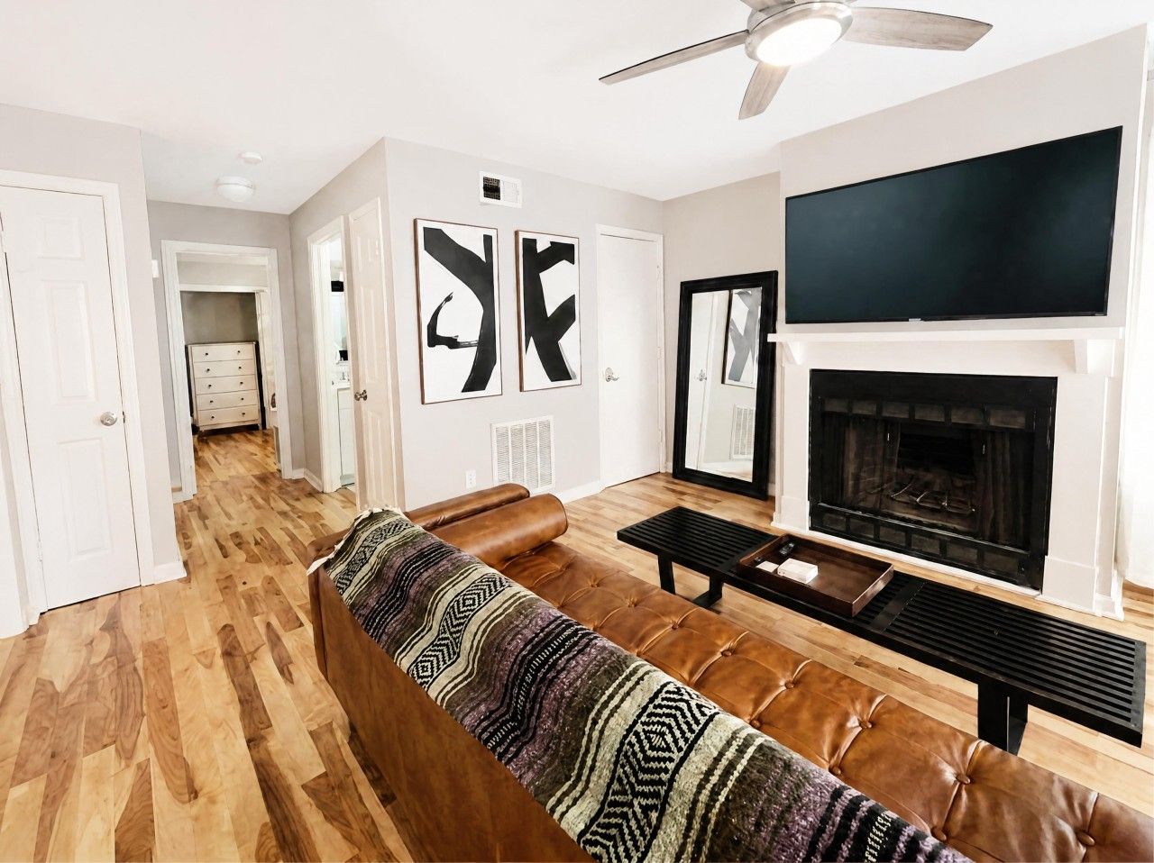 A living room with a brown leather sofa, a flat-screen TV mounted above a fireplace, and a ceiling fan.