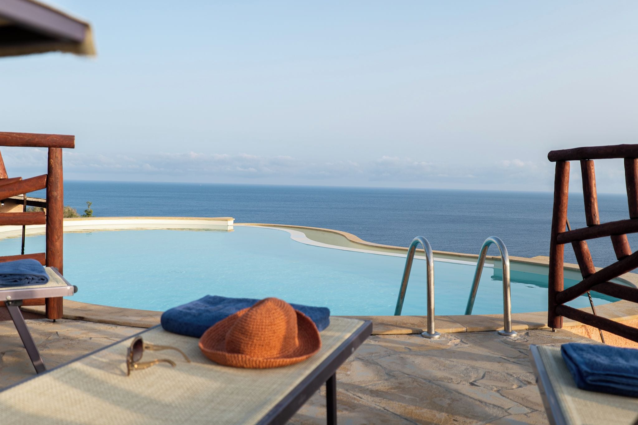 An infinity pool with a view of the ocean, wooden lounge chairs, and a hat on a table.