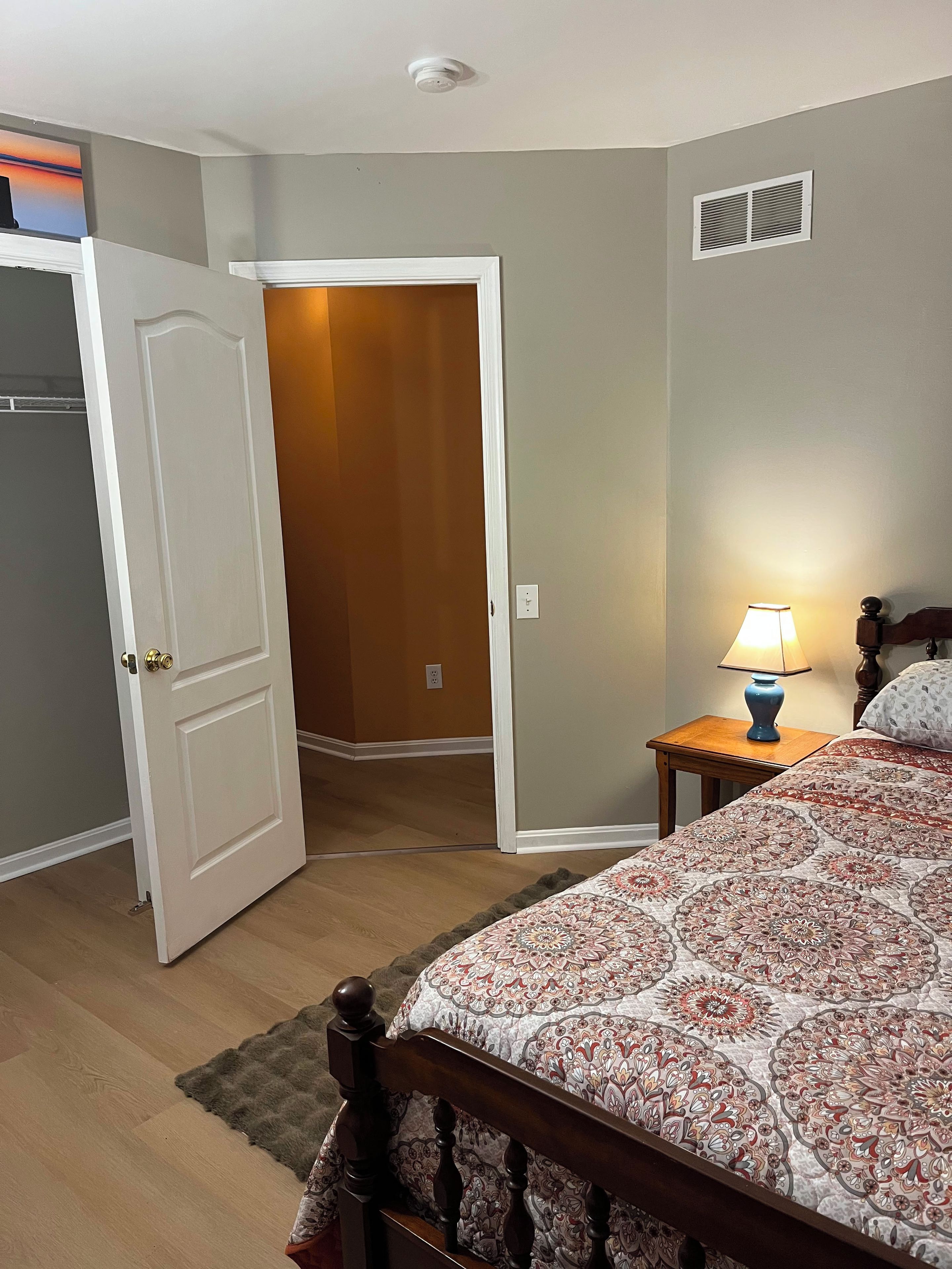 A bedroom with a bed, a nightstand with a lamp, a door leading to another room, and a wall-mounted vent.