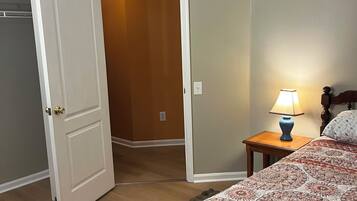 A bedroom with a bed, a nightstand with a lamp, a door leading to another room, and a wall-mounted vent.