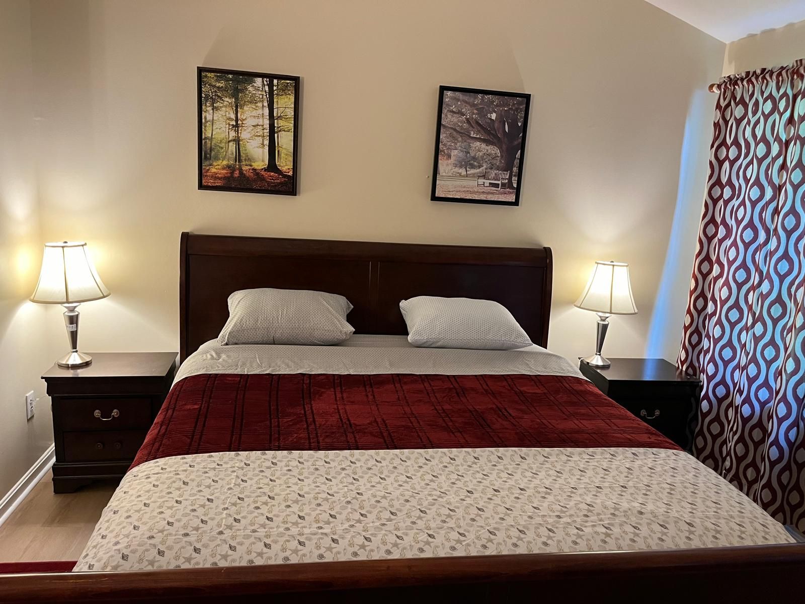 Un dormitorio con cama, dos mesitas de noche, dos lámparas, una manta roja, una colcha estampada y dos cuadros en la pared.