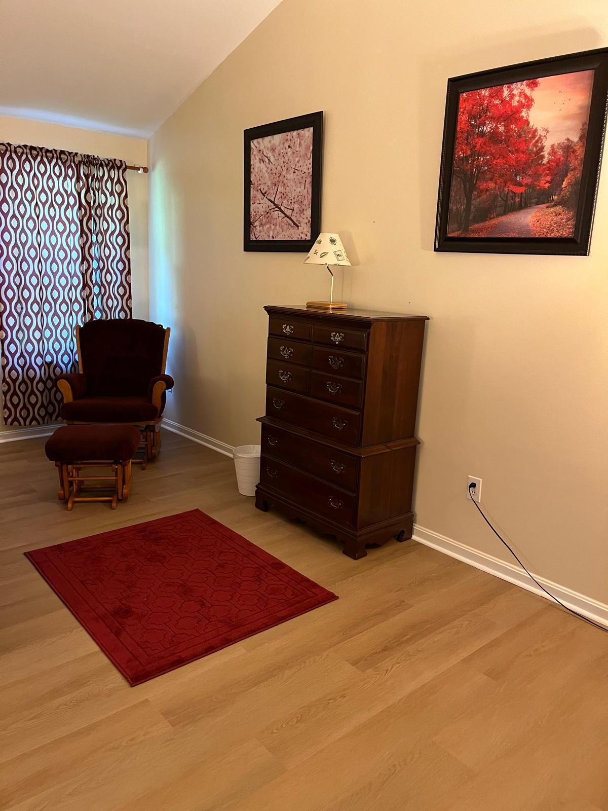 Una habitación con suelo de madera, un tapete rojo, un aparador de madera, una silla y dos cuadros en la pared.