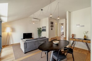 Ein modernes Wohnzimmer mit einem grauen Sofa, einem Esstisch und einem Fernseher. Es gibt Pendelleuchten und eine Pflanze.